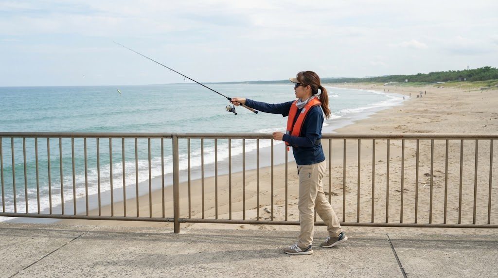 砂浜に面した低い堤防の柵の内側で、ライフジャケットを着た人が仕掛けを前方へ軽く投げ入れている様子