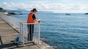 夏の朝の堤防で、白い安全柵の内側に立つライフジャケット姿の人が、サビキ用の竿で足元の海を見ている様子