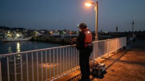 夕暮れの漁港の常夜灯の下で、ライフジャケットとヘッドライトを着けた人がアジング用の竿を構えている様子